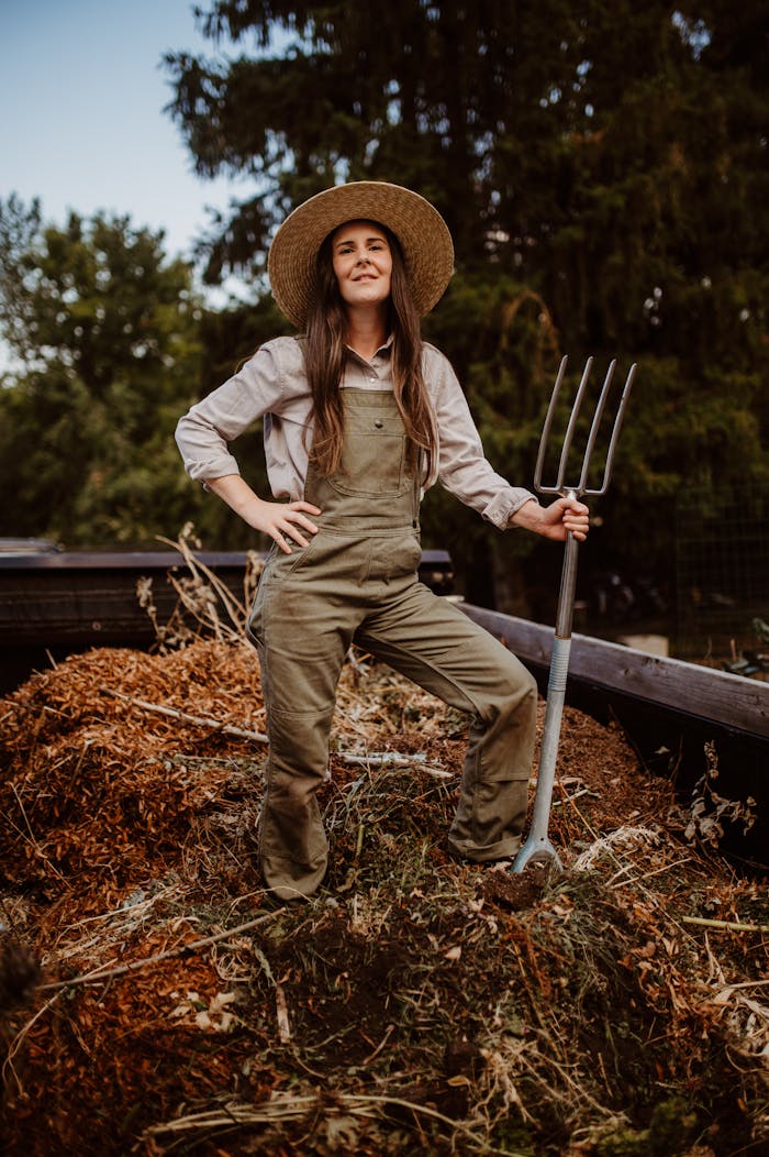 our-vision Woman gardener proudly standing with pitchfork by a compost heap outdoors.